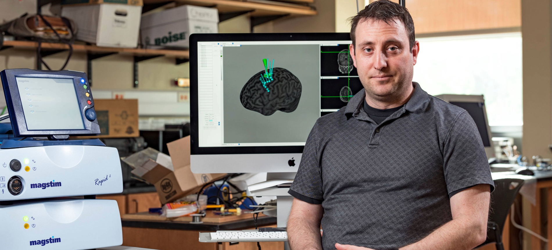 Lee Baugh smiling in front of a monitor showing brain research.