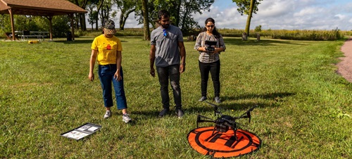 3 students operating a drone about to liftoff outside during the day.