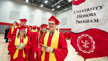 Students stand in a line wearing red graduation gowns. They stand next to a flag that says University Honors Program.