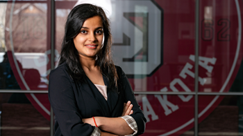 Priyam Pandey stands with her arms crossed in front of a large University of South Dakota logo.