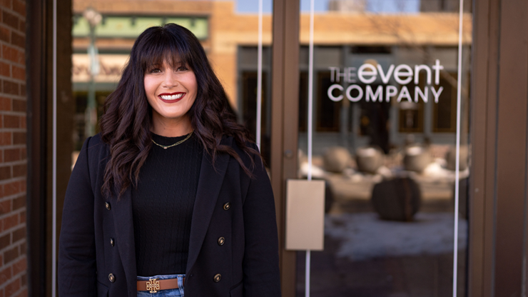 Addie Graham-Kramer smiles as she stands in front of glass doors with the words "The Event Company" on them.