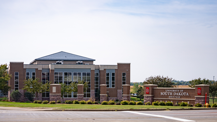 A street view of the USD – Sioux Falls campus that shows a building and a sign that says University of South Dakota Sioux Falls. 