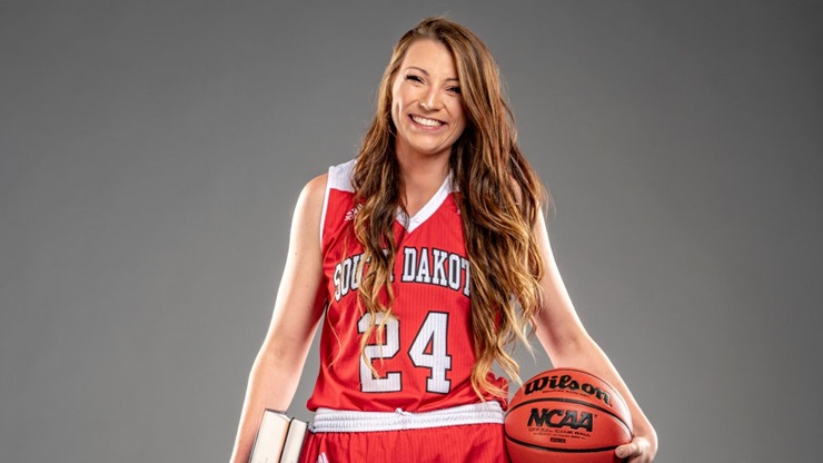 A portrait of Ciara Duffy in her USD basketball uniform. She holds books in her right hand and a basketball in her left hand. Her uniform is red, and she is number 24.