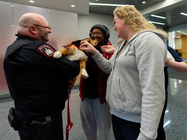 A police officer holds a corgi puppy while several students excitedly pet it.