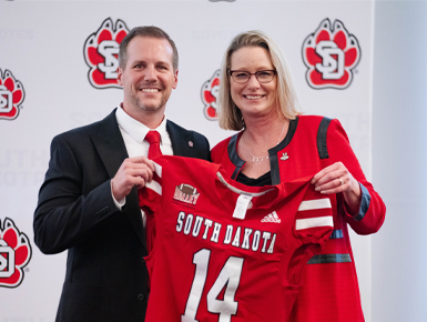 Jon Schemmel stands next to President Sheila K. Gestring. They hold up a number 14 football jersey and smile for a photo.