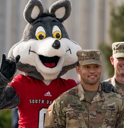 Charlie holding up a Yote hand symbol while standing behind a smiling USD military student in uniform.