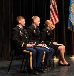 3 USD Military Graduates sitting in formal dress wear on stage.