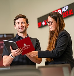A male and female USD student standing in the Ellis Finance Lab reading an open notebook.