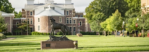Campus photo of Legacy coyote sculpture in the foreground and Old Main in the background