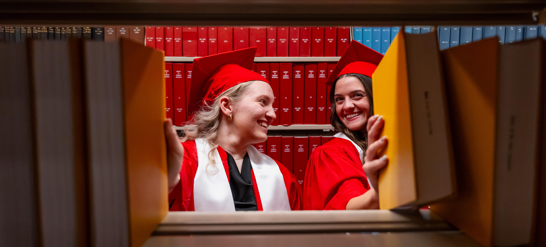 Two students in caps and gown grabbing books from a shelf