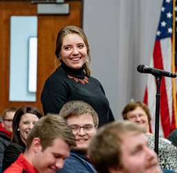 Photo of students all smiling in a large room with one standing in front of a microphone