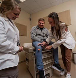 Two med students helping a patient sitting and smiling on the office table.