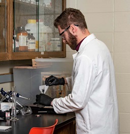 Male Researcher in White Lab Coat and Goggles Adding Powder to Beaker.