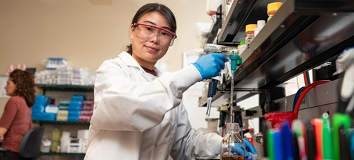 A BBS student holding scientific instruments in a lab and smiling at the camera.