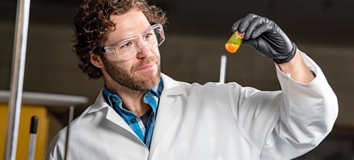 Male Researcher in White Lab Coat and Goggles Examining a Vial.