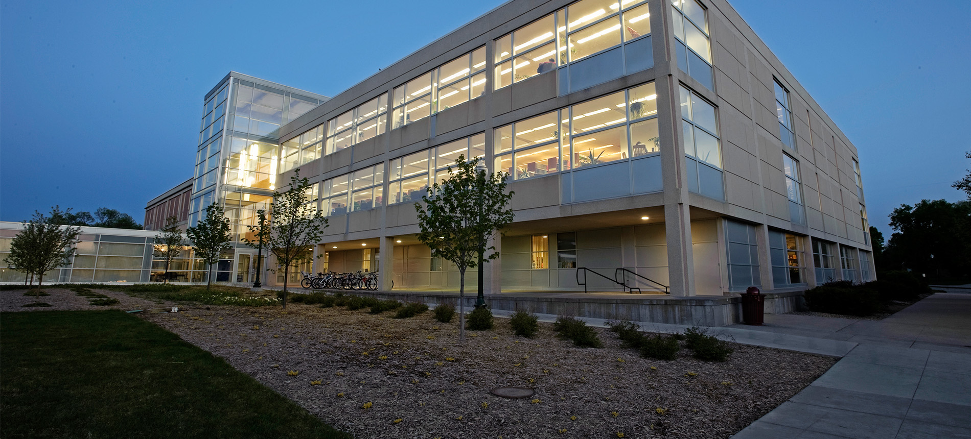 Photo of the outside of I.D. Weeks library in the early morning hours. Concrete and glass building with yellow lighting shining through the 3 story buildings many windows.