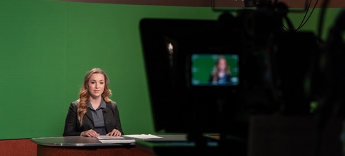 Student sitting as a news anchor in front of a green screen with a news camera on them