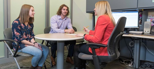 Female in Red Blazer Sitting at Table with Two Individuals.