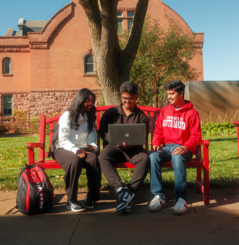 Three students sitting outside on a bench on USD Vermillion campus looking at one laptop.