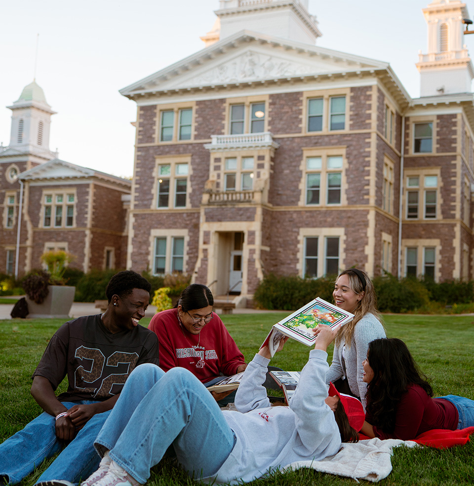 Five students laying on campus grass reading a book and laughing with eachother.