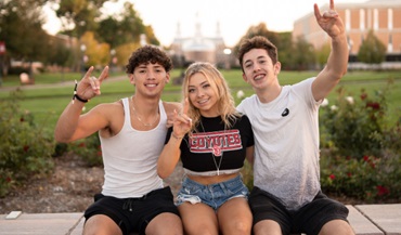 Three students sitting on the old main lawn smiling and posing with Coyote hand symbols.