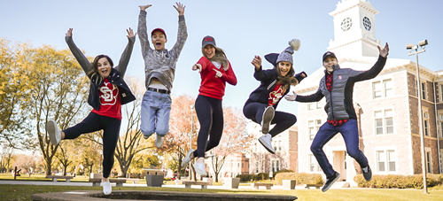 Photo of 5 students excited and all jumping at once in front of iconic Old Main