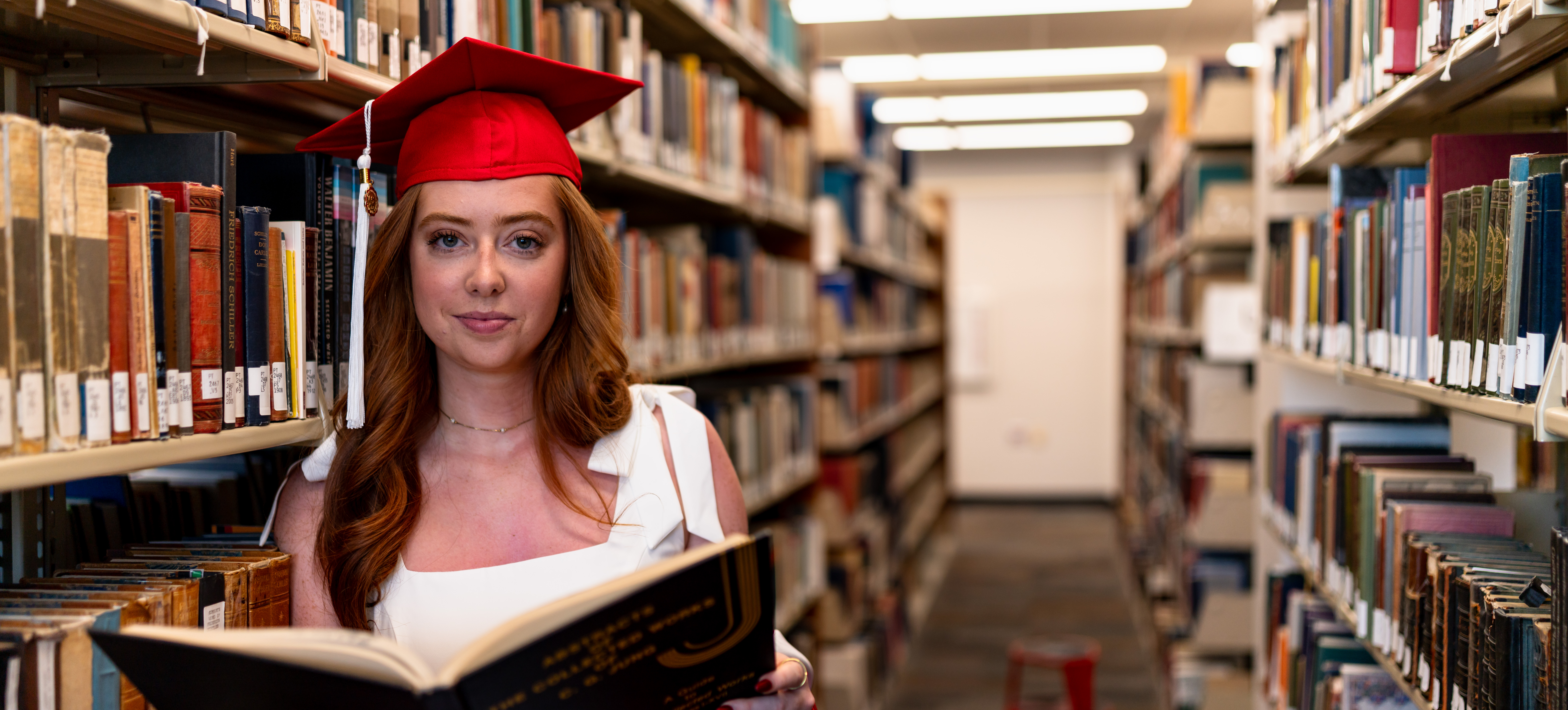 female student reading a book in the library in her graduation cap