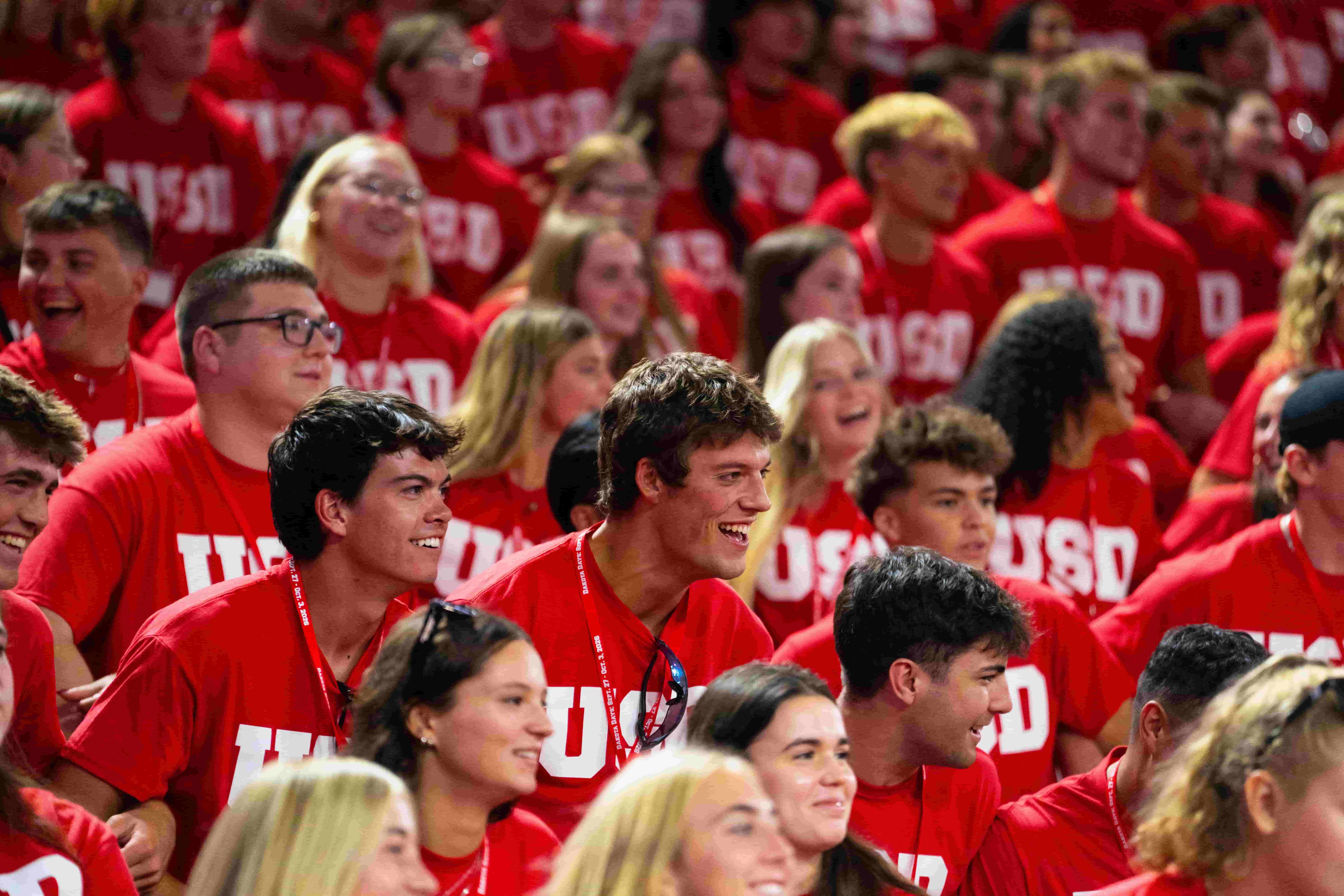 A packed crowd of smiling college students in red “USD” shirts cheer together at a spirited campus event, creating an energetic, celebratory atmosphere.