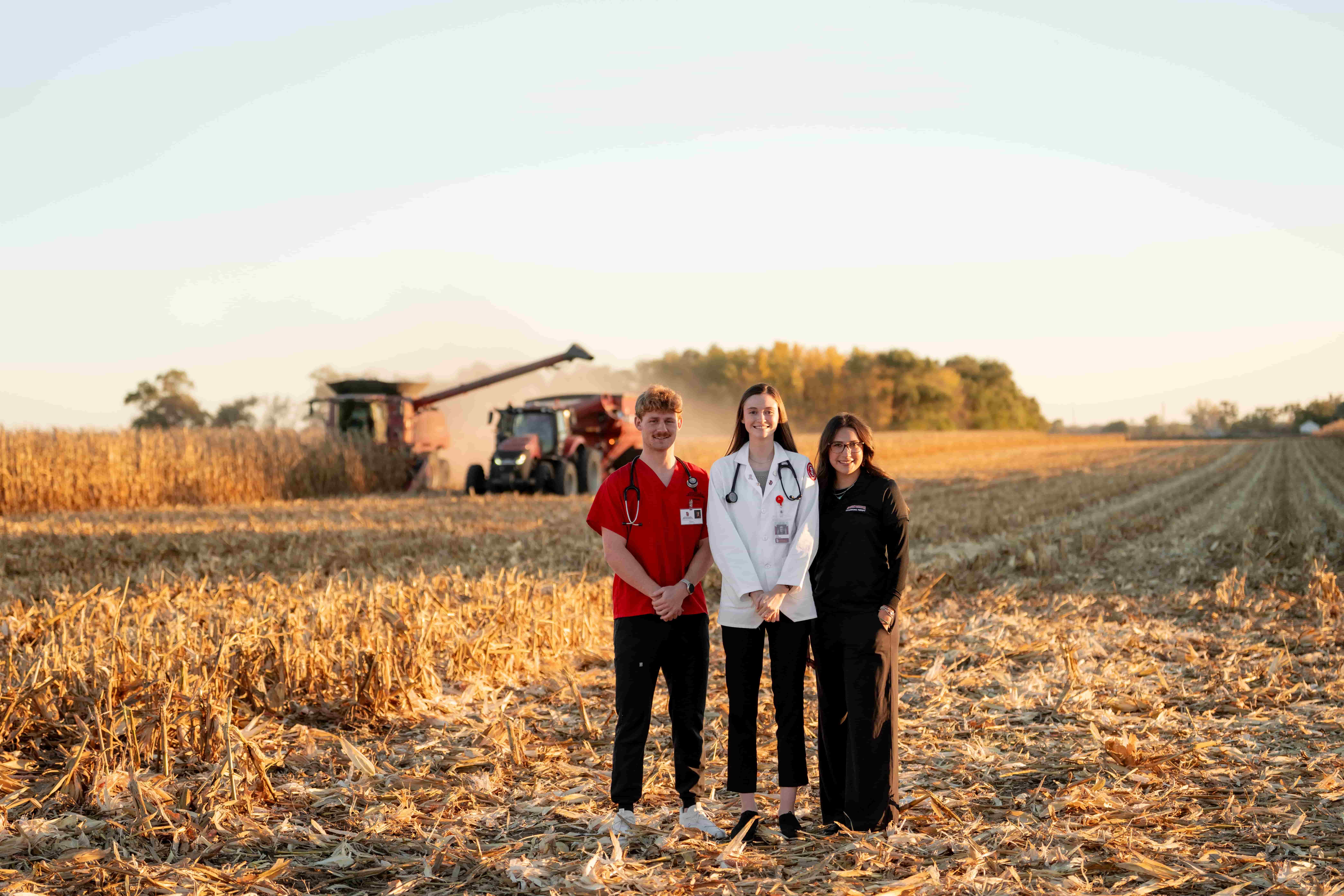 Three students stand in a harvested field at golden hour, conveying collaboration and a commitment to rural healthcare.