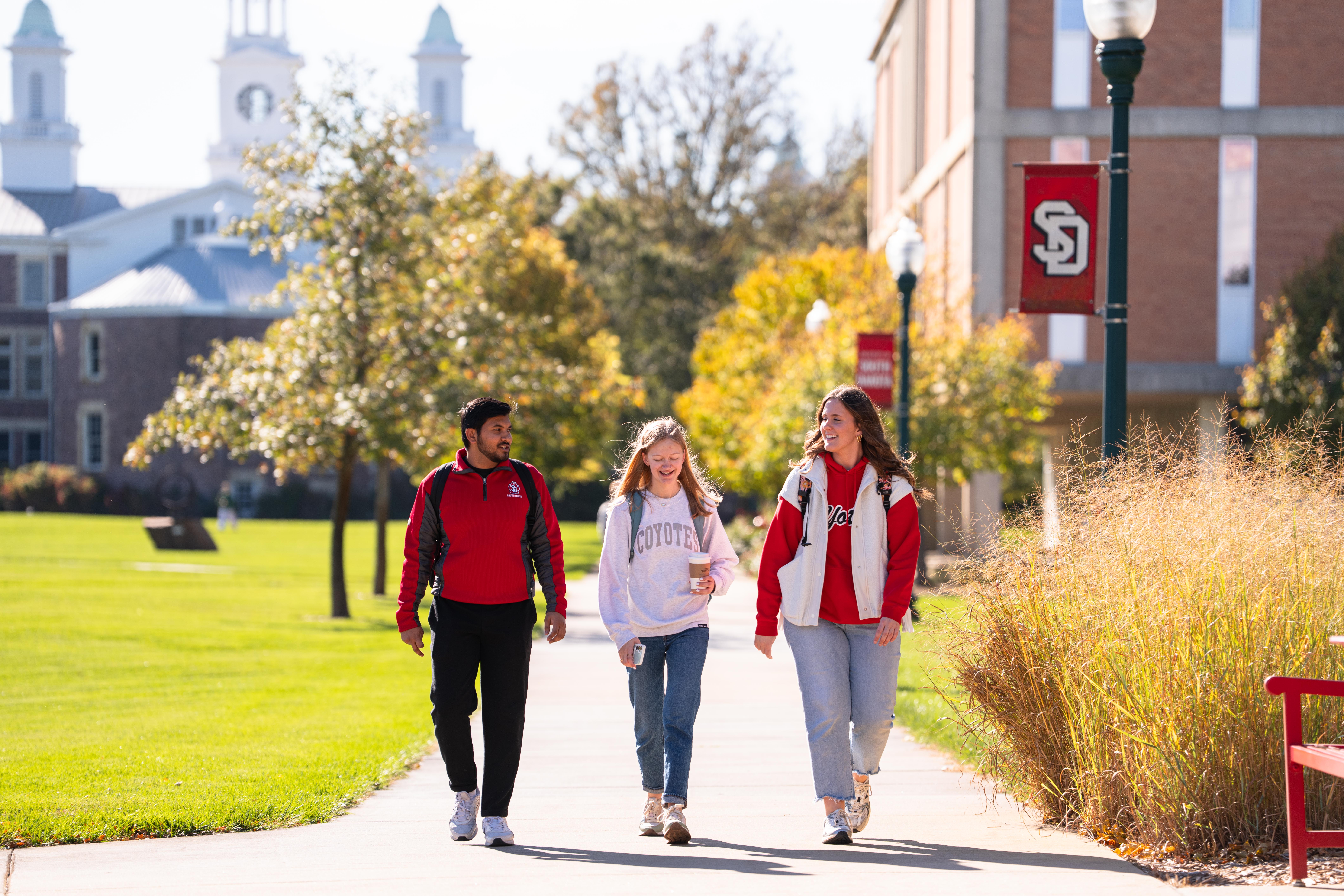 Three college students walk and chat on a sunny campus path, with golden autumn trees, historic buildings, and a red university banner.