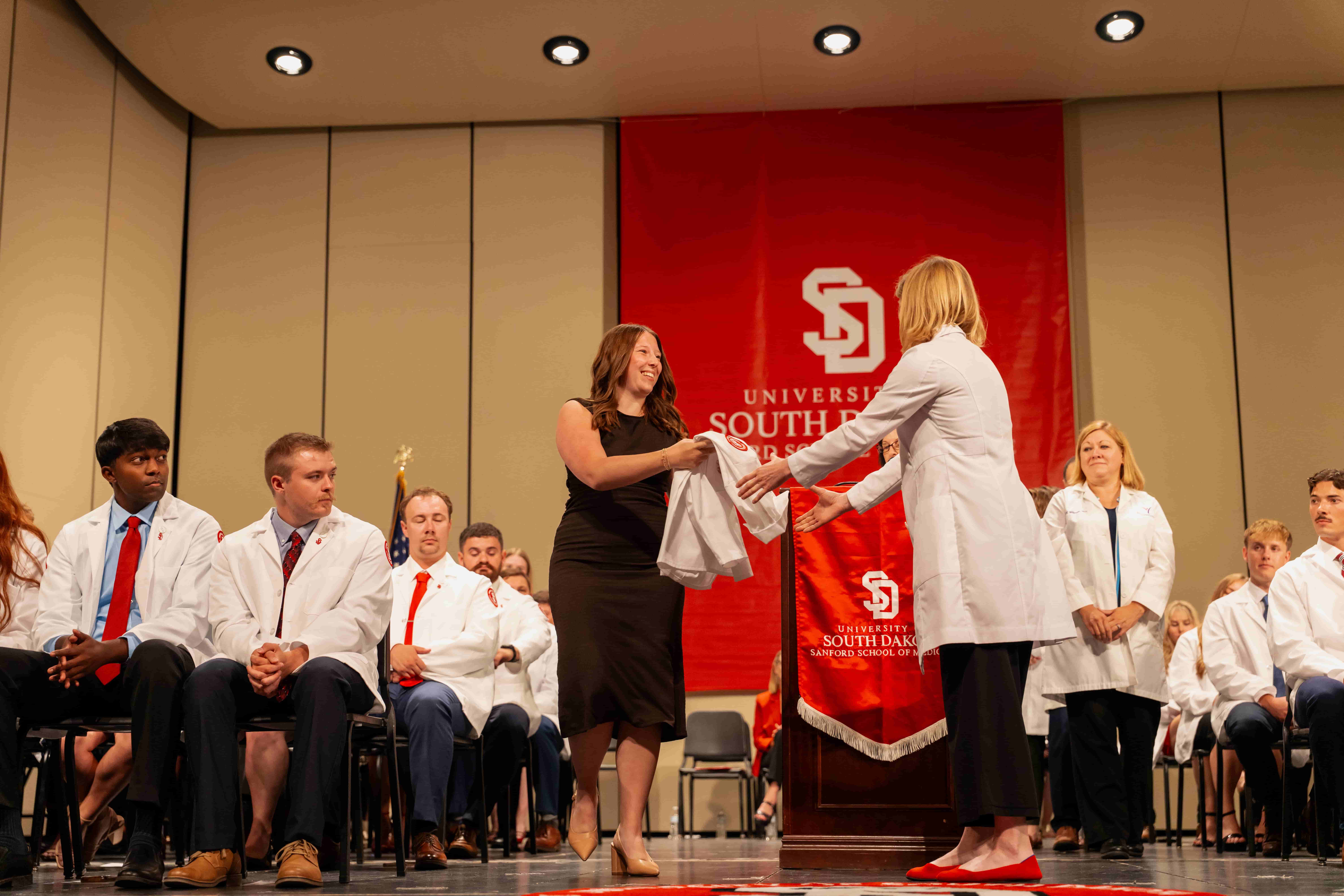 At a university ceremony, a smiling woman in black presents a folded garment to a woman in a white coat, as faculty and students watch onstage.