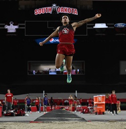 A USD men's track long jumper in mid air during a competition.