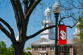 A picture of a USD Banner on campus during the spring with Old Main in the background.