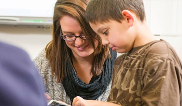 A teacher assisting a young student while both are smiling in a classroom.