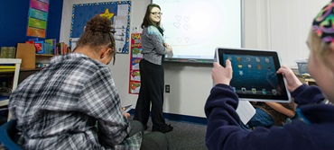 A teacher at a whiteboard while a student holds up an I pad.
