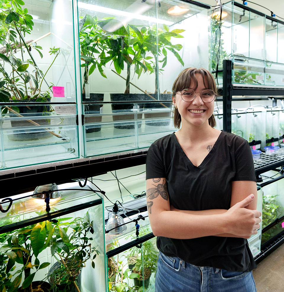 A student standing with their arms crossed in front of glass enclosures for chameleons