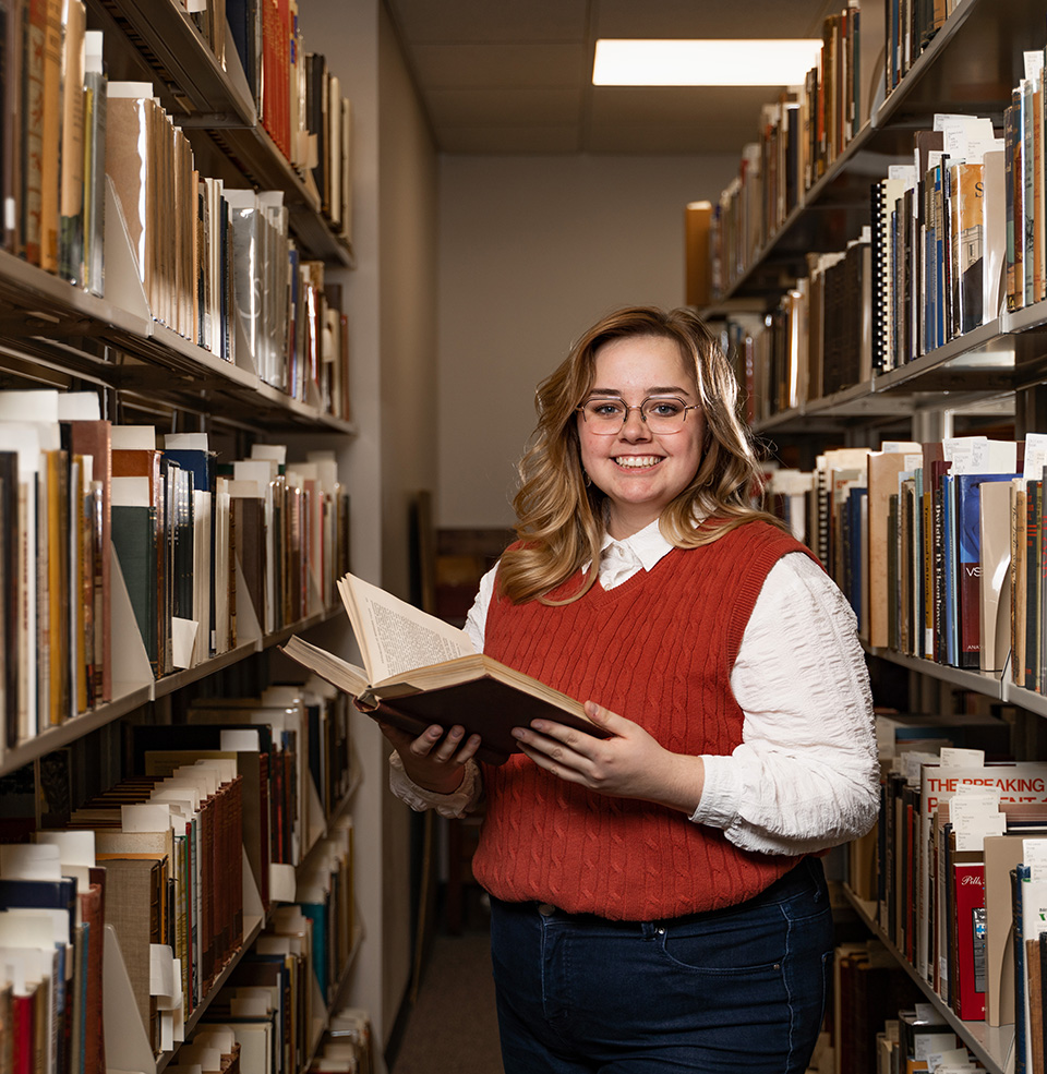 A student standing in between aisles of books in the library