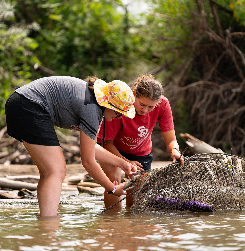 Two students standing knee deep in the missouri river with a fishing net and holding a fish