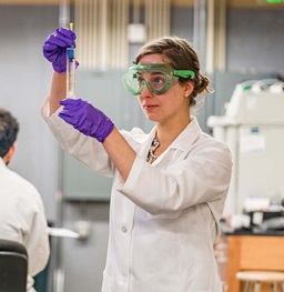 Female Researcher in White Lab Coat and Goggles Examining a Tube
