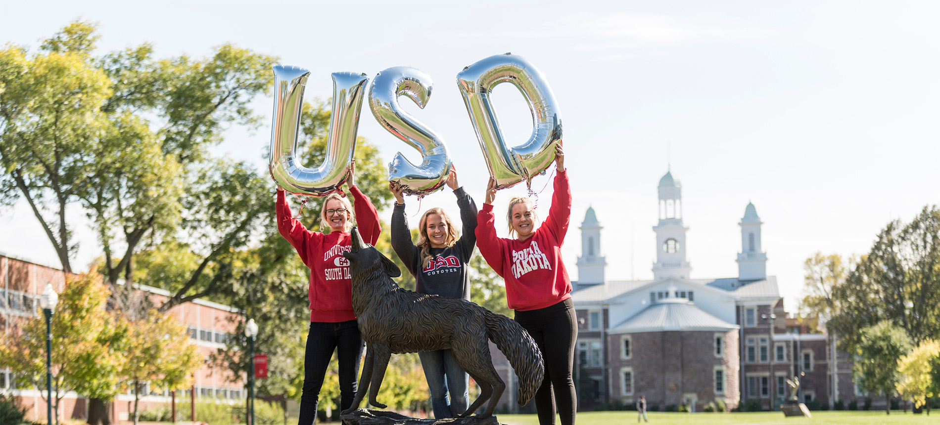 Students standing in front of old main holding giant U S D balloon over their heads