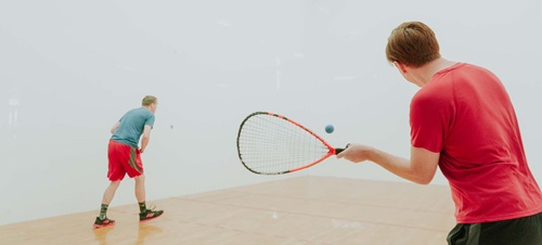 Two male students playing racquetball on a hardwood court.