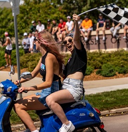 A sorority girl waving a flag riding on the back of a moped during bid day.