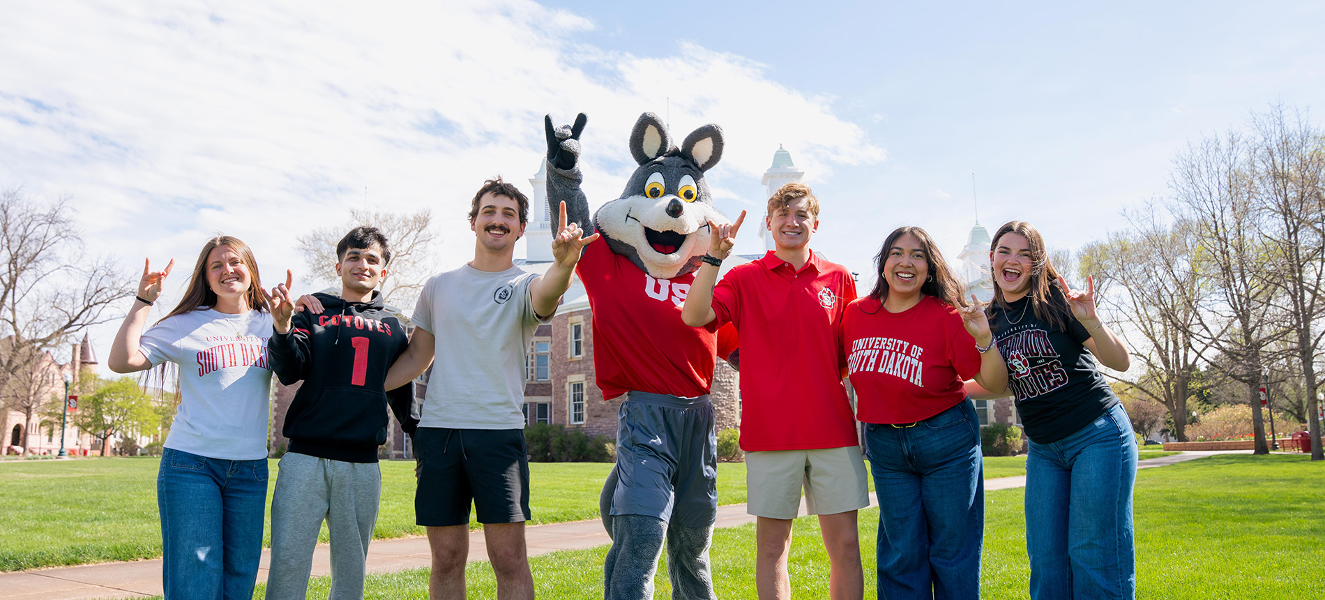 Charlie standing with students in front of Old Main holding up a hand coyote sign