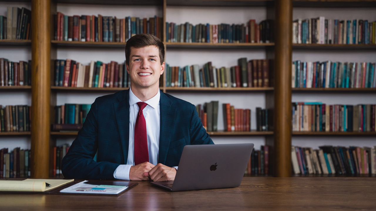 A photo of Trenton Hoekstra sitting at a table with his laptop. There is a bookshelf behind him.