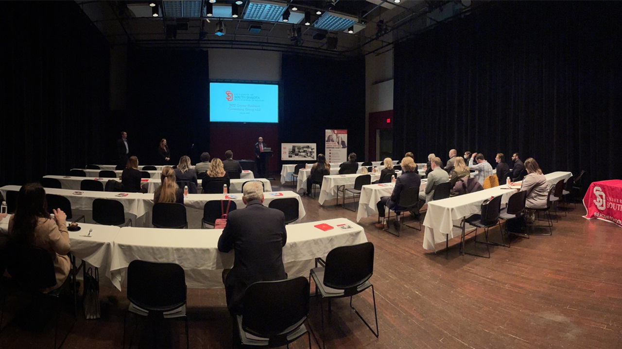 Business professionals sit at tables in a dark room and look at a presentation.