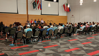 Rows of students sit in chairs and listen to five speakers on stage.