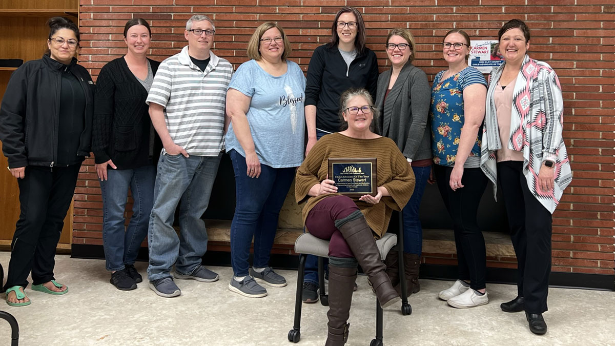 Carmen Stewart sits on a chair and holds an award plaque. Eight people stand behind her.