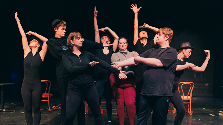 Musical theatre students stand in a group and spread out their arms on stage.