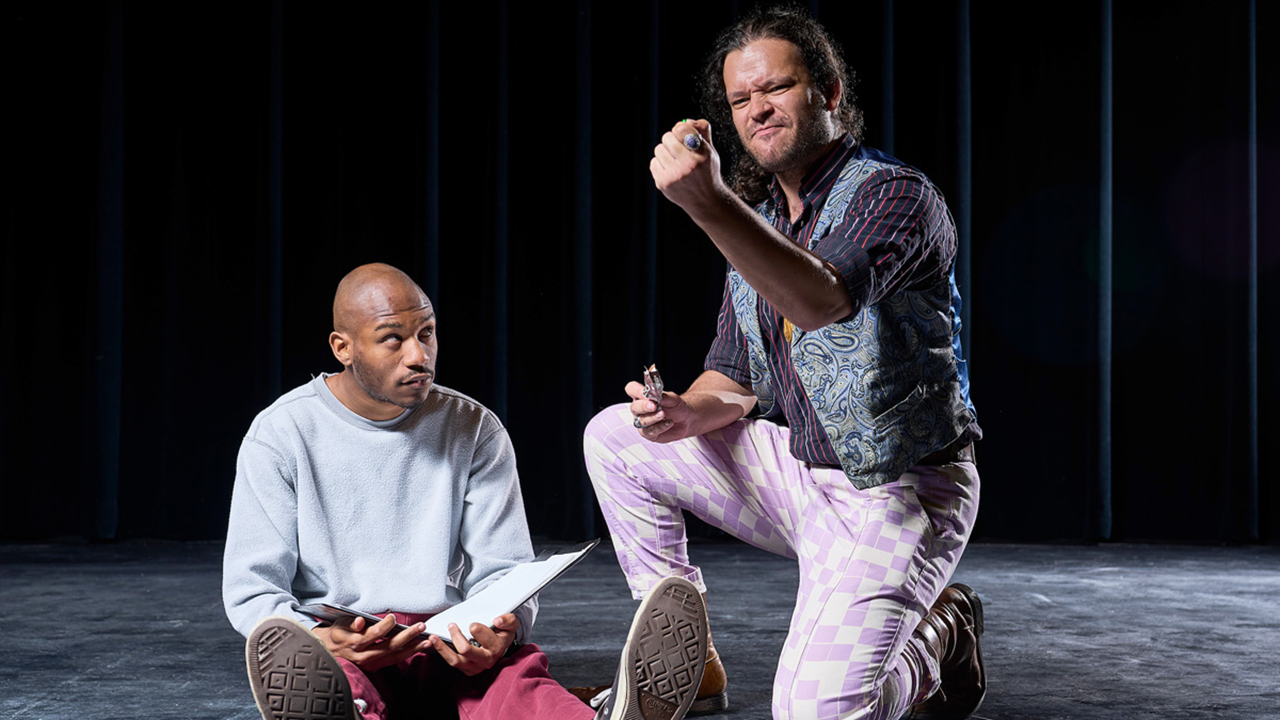 Two actors sit on a dark stage. One sits with his legs out in front of him, and the other sits on one knee while holding up a fisted hand.