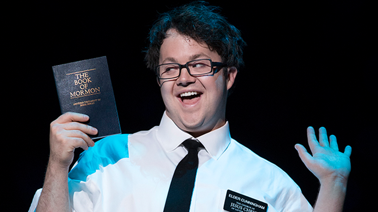 A picture of Cody Jamison Strand with a spotlight on him. He has his hands up and is holding a booklet that says The Book of Mormon. He is wearing a white shirt with a black tie and has a nametag that says Elder Cunningham.